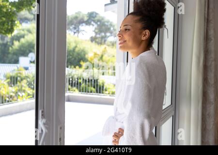 Mixed race woman wearing bathrobe standing by terrace door and smiling Stock Photo