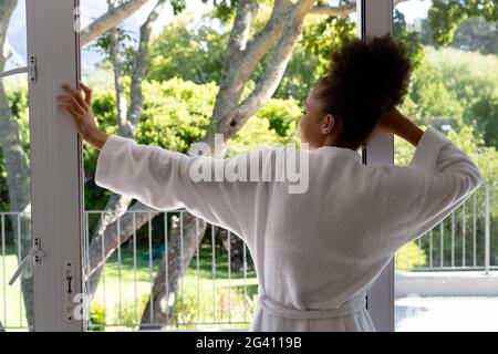 Mixed race woman wearing bathrobe standing by terrace door and smiling Stock Photo