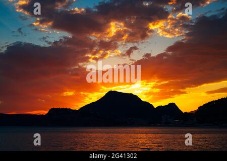 Bright fiery sunset behind the silhouette of a mountain over the sea ...