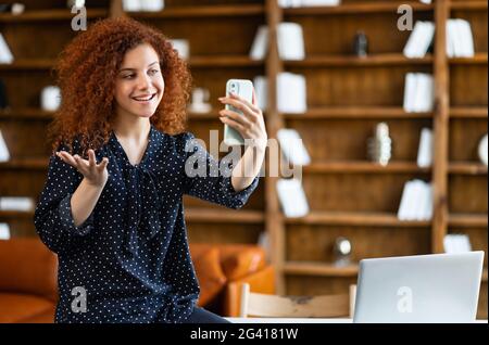 Video connection on the distance. Positive young curly woman using the smartphone for video call. Smiling red-haired female office worker has pleasant online meeting sitting in modern office space Stock Photo