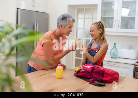 Senior grandmother giving granddaughter packed lunch and lollipop in kitchen Stock Photo