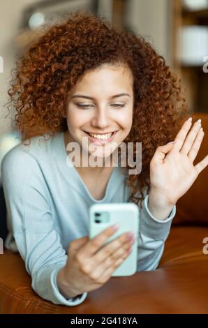 Happy curly red-haired woman resting at home on leather sofa, talking chatting via video call, using app on smartphone, has conversation with family, greeting friends, waving hand, vertical portrait Stock Photo