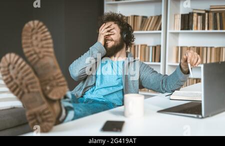 Facepalm. A young programmer works remotely at home. A young man smiles, covering his face with his hands sitting at a table in Stock Photo