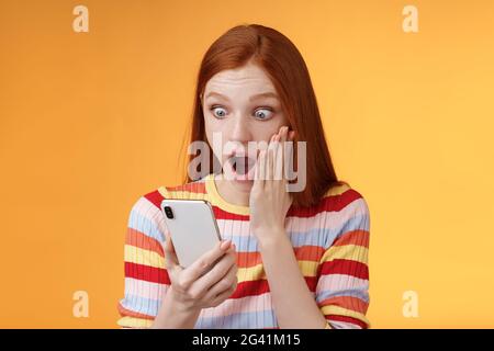 Omg what heck. Portrait shocked concerned young redhead sensitive impressed redhead woman stare smartphone display touch cheek d Stock Photo