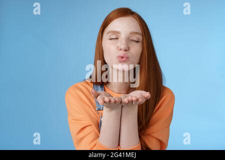 Passionate dreamy pretty redhead girl sending air kisses camera close eyes folding lips hold hands near mouth give muah internet Stock Photo