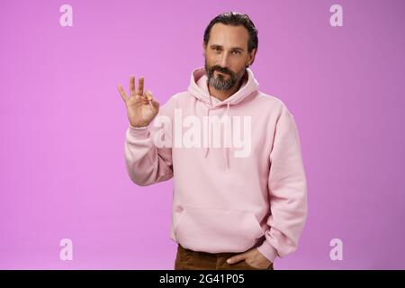 Confident good-looking lucky old macho guy bearded grey hair in stylish pink hoodie hold hand pocket show okay ok approval gestu Stock Photo