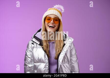 Stylish friendly charismatic blond woman in silver shiny jacket hat sunglasses ready learn snowboarding smiling laughing happily Stock Photo
