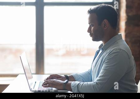 Side view at confident indian businesswoman using laptop in the office. Young male mixed-race employee in casual wear typing and chatting online, develops software indoors Stock Photo