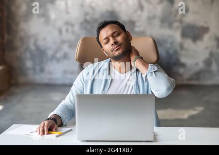 Tired indian freelancer guy massaging back of the neck, feels muscle tense, young hispanic man take a break from work with a laptop sitting in the home office with eyes closed, suffers from back ache Stock Photo