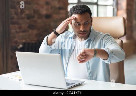 Confused indian man in smart casual shirt using the laptop in loft style office, hurry up to meet the deadline, watching on his smart watch and checking time Stock Photo