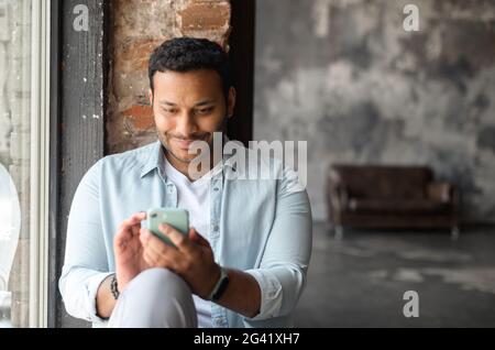 Happy and smiling indian dark-haired man in casual shirt using smartphone in loft style apartment, sits on windowsill texting, chatting online, web surfing, using new mobile app in rested atmosphere Stock Photo