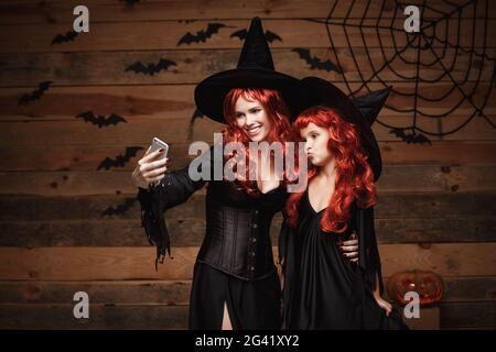 Halloween Concept - Beautiful caucasian mother and her daughter with long red hair in witch costumes taking a selfie with smartp Stock Photo