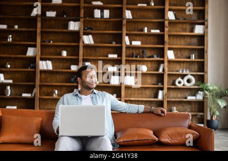 Cheerful indian man wearing casual shirt sitting down on the sofa and using laptop at modern home office, confident male entrepreneur looks away and smiles, a book shelves on background Stock Photo