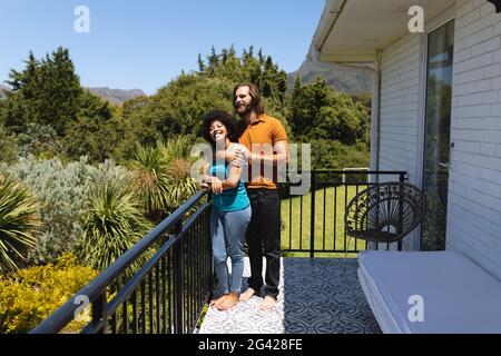 Happy diverse couple standing on balcony embracing and smiling Stock Photo
