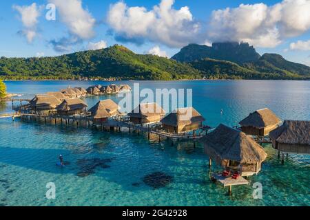 Aerial view of man on SUP stand up paddle board in Bora Bora lagoon with overwater bungalows of Sofitel Bora Bora Private Island Resort behind, Vaitap Stock Photo
