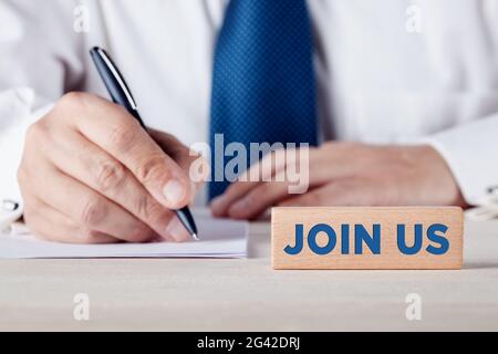 Join us message written on a wooden block with a businessman writing on a sheet of paper at the background. Membership registration concept. Stock Photo