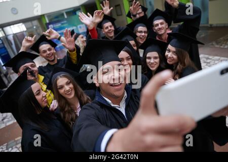 Group of happy international students in mortar boards and bachelor gowns with diplomas taking selfie by smartphone Stock Photo