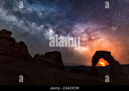 The Pack Creek Fire in the La Sal Mountains lights up the night sky in the distance, with the Milky Way over Delicate Arch in Arches National Park, Ut