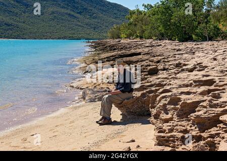 Cape Gloucester, Queensland, Australia - June 2021: Active male pensioner sitting on a rocky coastal shore holding the controls of a drone as he learn Stock Photo