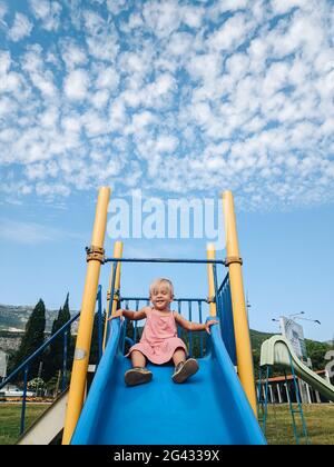 A little girl is sliding down a children's slide against the background of a blue sky with white clouds. Stock Photo