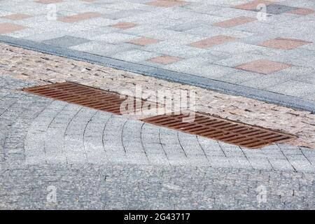 2 rusty iron hatch rectangular grating of the drainage system on the road with paved stone tiles near the pedestrian sidewalk made of granite close-up Stock Photo