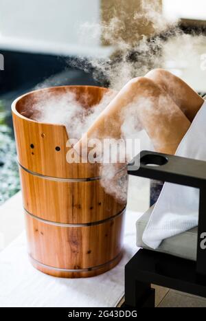 A woman having a footbath, in wooden barrel , with steam billowing. Horizontal. Kuala Lumpur, Malaysia. Stock Photo