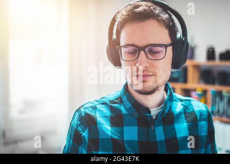Enjoying music at home: Young Caucasian man is listening to music with headphones. Blue checked shirt. Stock Photo