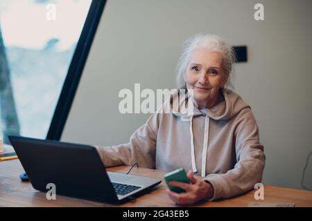 Aged elderly woman with laptop and mobile phone working on computer at table Stock Photo