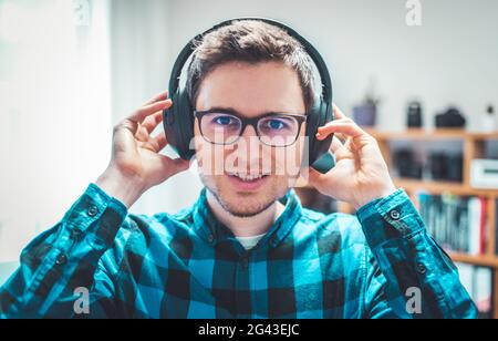 Enjoying music at home: Young Caucasian man is listening to music with headphones. Blue checked shirt. Stock Photo