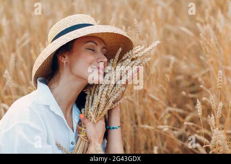 Young Woman in straw hat holding sheaf of wheat ears at agricultural field Stock Photo