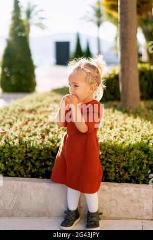 Pretty little girl in a terracotta colored dress and white tights is snacking on a pie in a park on a sunny day Stock Photo