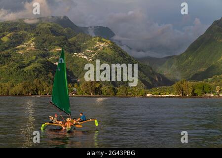 People enjoy a trip on an outrigger canoe with sails in front of a lush mountain backdrop, near Papeete, Tahiti, Windward Islands, French Polynesia, S Stock Photo
