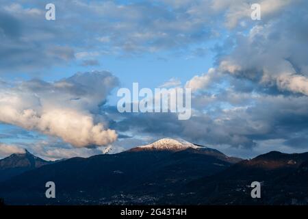 Snow-covered peak of the mountain is illuminated by the sun, the velvet white clouds on the blue sky, on Lake Como in Italy. Stock Photo