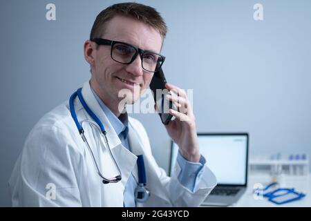 Smiling doctor having phone call at desk. Physicianr talking with his patient in office clinic sitting at table with laptop. Ger Stock Photo