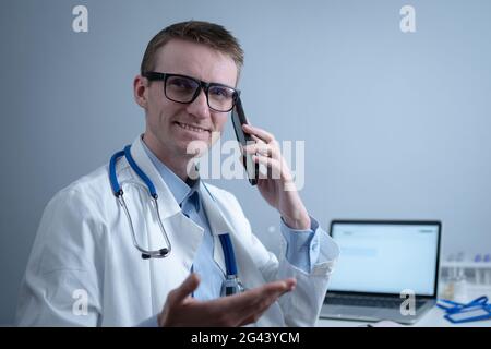 European doctor in white medical coat and glasses consults patient on cell phone in clinic office, sitting at table with laptop. Stock Photo