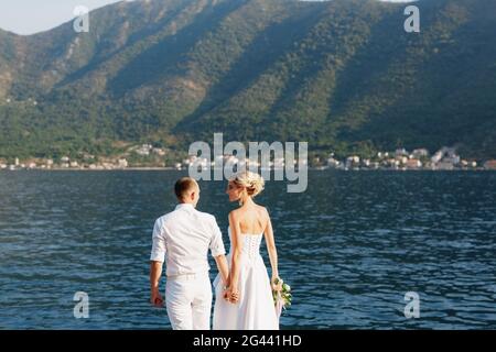 The bride and groom stand holding hands on the shore of the Bay of Kotor, back view Stock Photo