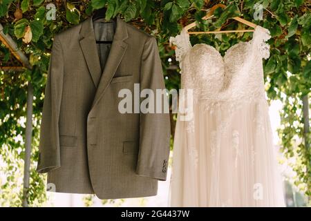 Close-up of the bride's white wedding dress and the groom's gray jacket on hangers on the arch with green leaves in the yard. Stock Photo