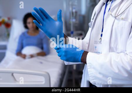 Midsection of african american male doctor putting on gloves, woman in hospital bed in background Stock Photo