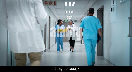 Diverse couple of female doctors walking through hospital corridor discussing Stock Photo