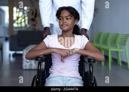 African american male doctor pushing smiling mixed race girl in wheelchair through hospital corridor Stock Photo