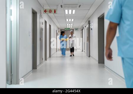 Diverse couple of female doctors walking through hospital corridor discussing Stock Photo