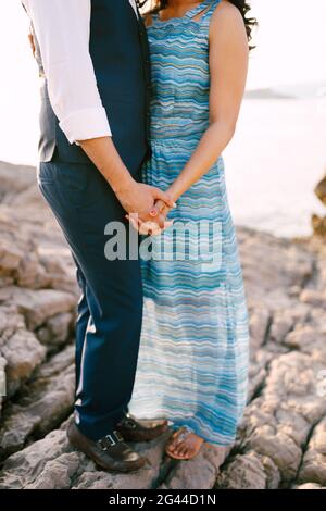 Man in a suit and woman in a long dress with an ornament stand on a stone shore holding hands Stock Photo