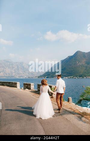 The bride and groom walk hand in hand along the road along the coast in the Bay of Kotor near Perast, back view Stock Photo