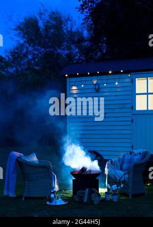 Nightshot of bonfire, set against an old wooden bungalow, arranged with wicker chairs and blankets, Bray, United Kingdom Stock Photo