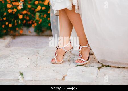 Bride's legs in stylish sandals with crystals with high heels peek out from under the wedding dress, close-up Stock Photo