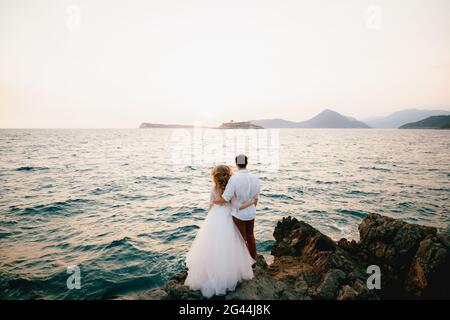 The bride and groom stand hugging on the rocks by the sea and look into the distance Stock Photo