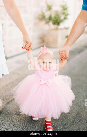 A charming little girl in a pink dress, sneakers and with a crown on her head holds mom and dad by the hand Stock Photo