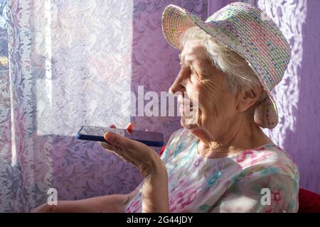 Charming elderly woman in rectangular black rimmed eyewear using voice recognition function on cell phone. Modern senior female holding electronic gadget, sending vocal message via online messenger Stock Photo
