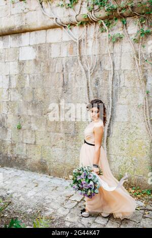 Beautiful bride in a pastel dress and high-heeled shoes walks with a luxurious bouquet of flowers along a stone wall Stock Photo