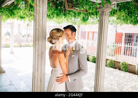 The bride and groom stand between the columns, hugging and smiling. The bride put her hands on the groom's chest. Stock Photo